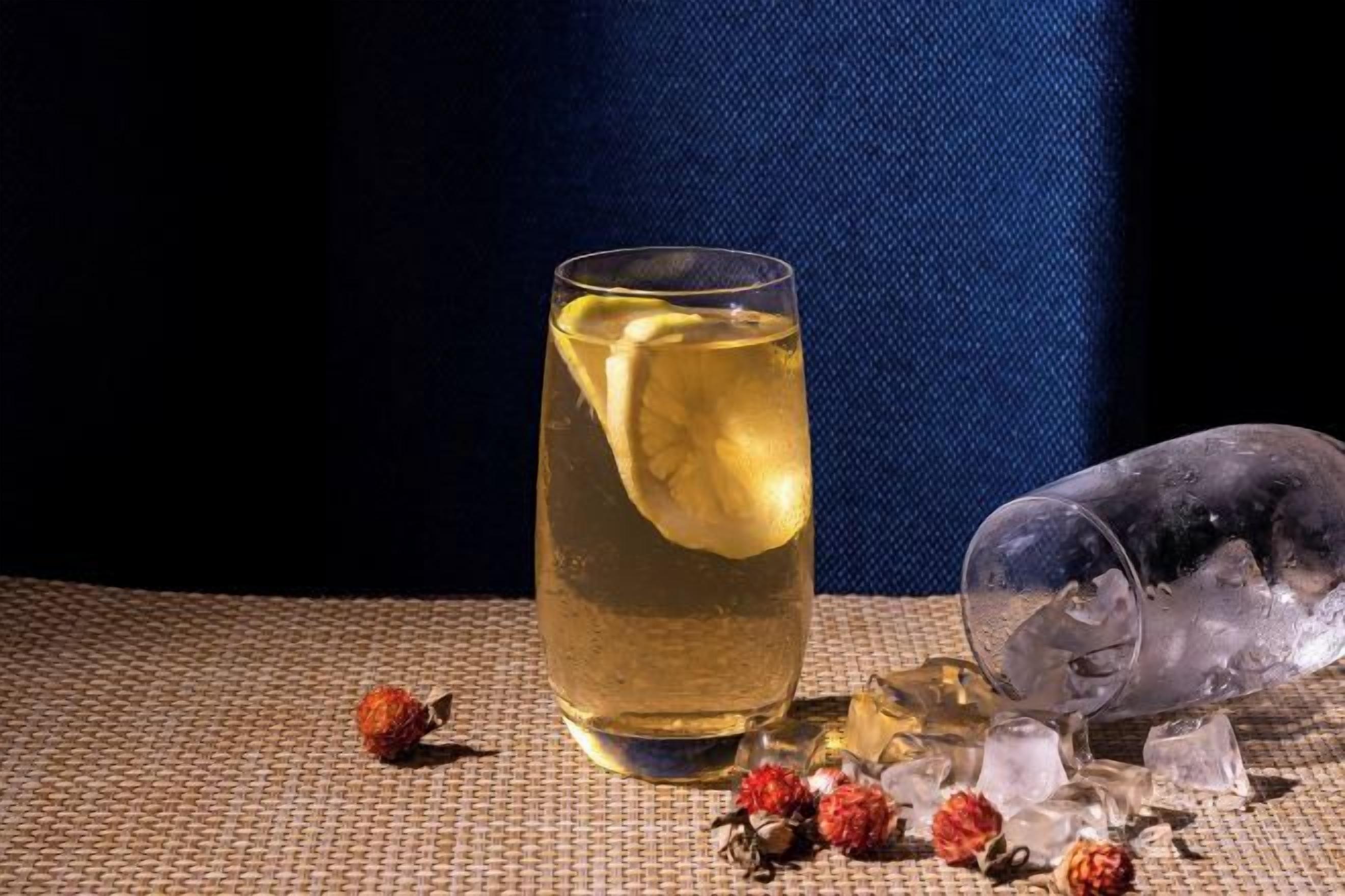 A glass of amber-colored liquid with a lemon slice, ice cubes, and scattered berries on a textured surface.