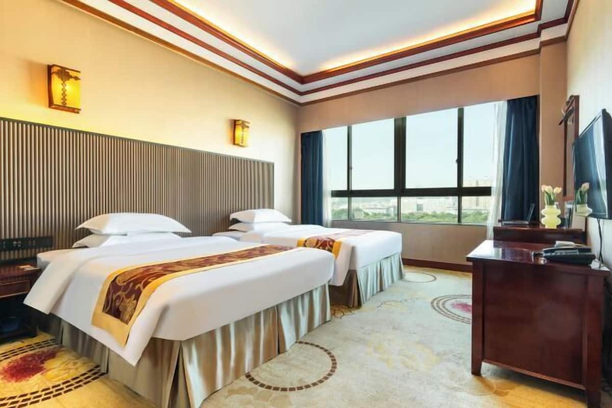 A hotel room with two beds, a desk, a TV, and a large window offering a city view.