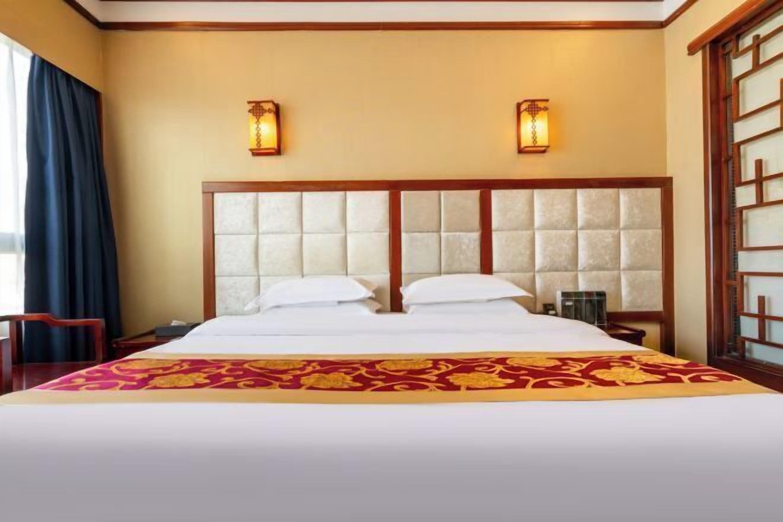A hotel room with a large bed, two wall sconces, a wooden headboard, and a decorative bed runner.