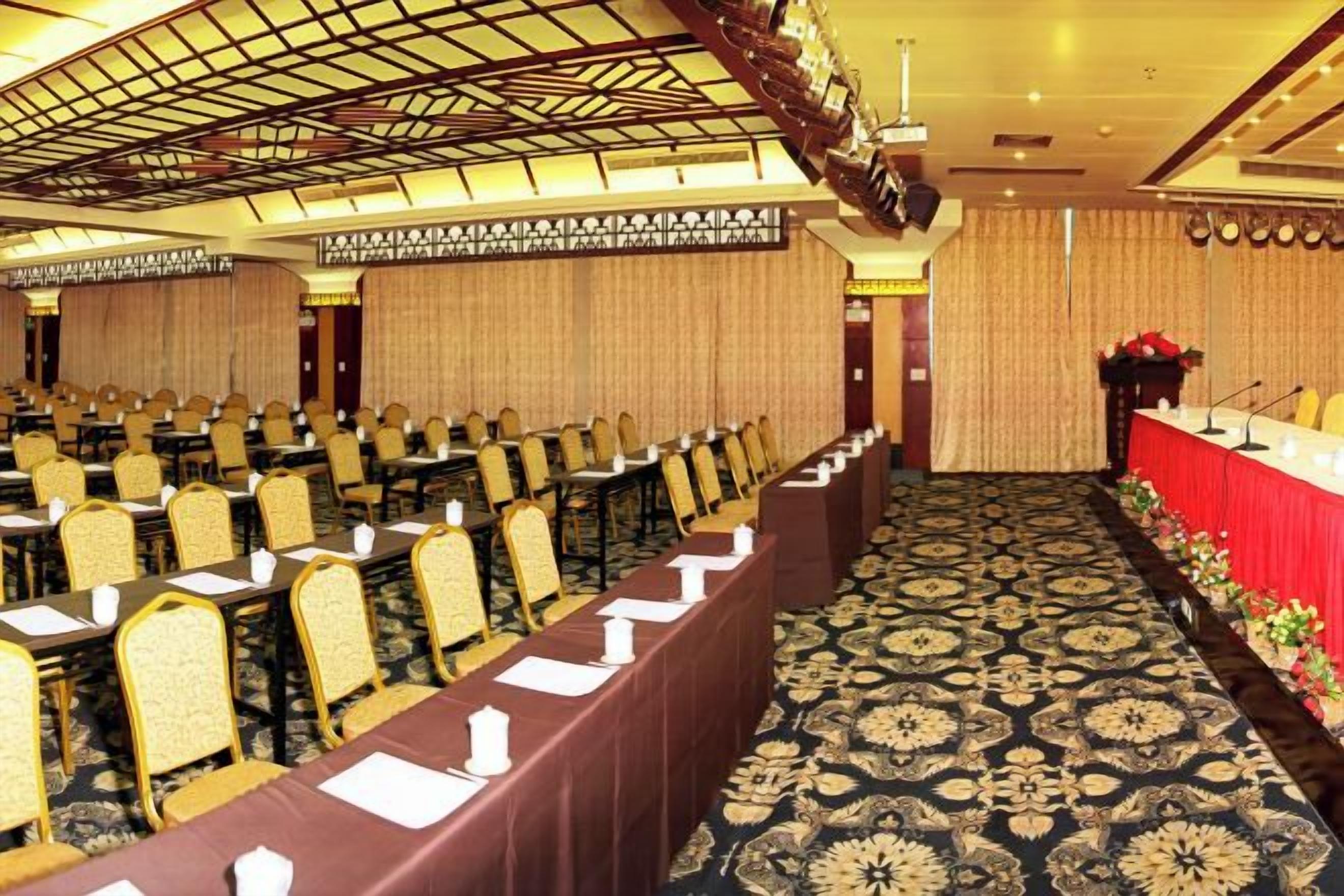 A conference room with rows of tables and chairs, a stage with a red tablecloth, and a patterned carpet.