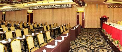 A conference room with rows of tables and chairs, a stage with a red tablecloth, and a patterned carpet.