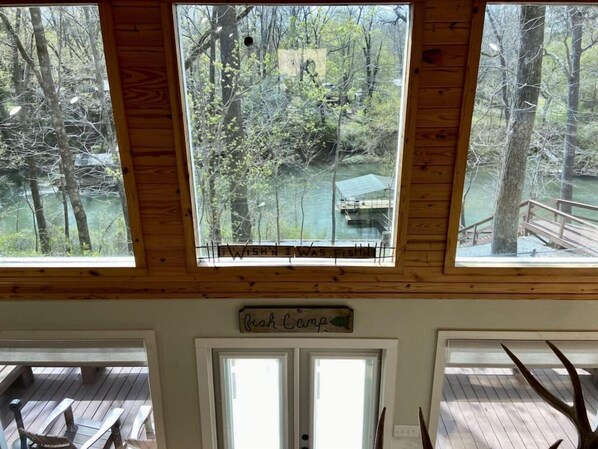 A cabin with three large windows offering views of a forest and a dock. A sign above the door reads "Bush Camp."