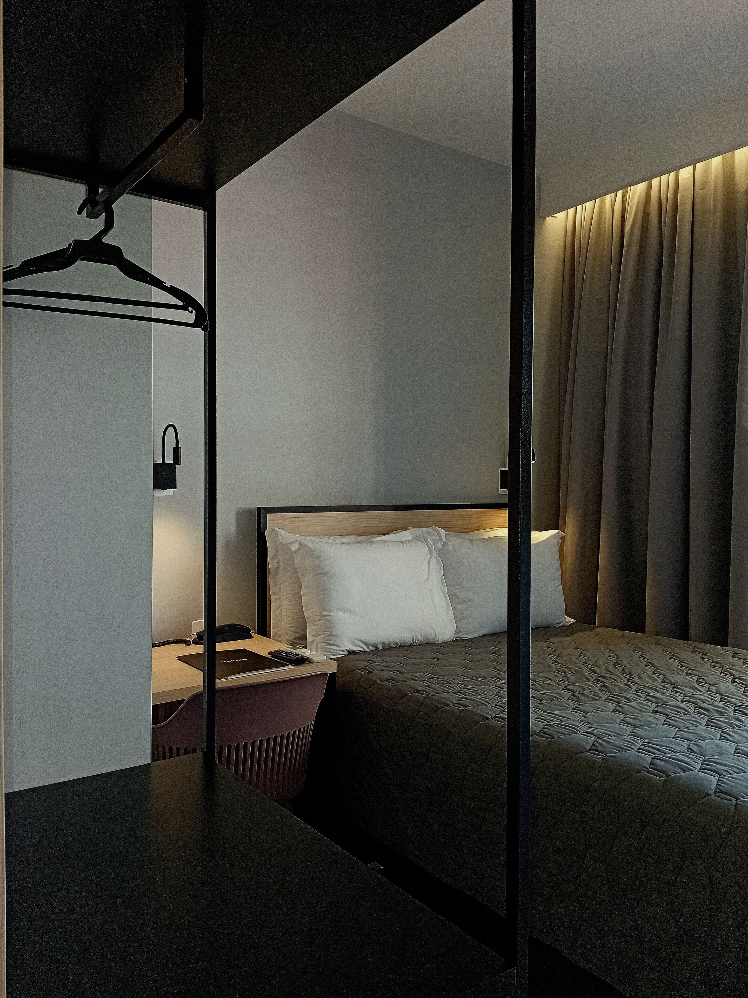 A hotel room with a bed, bedside table, lamp, and a hanging coat rack.