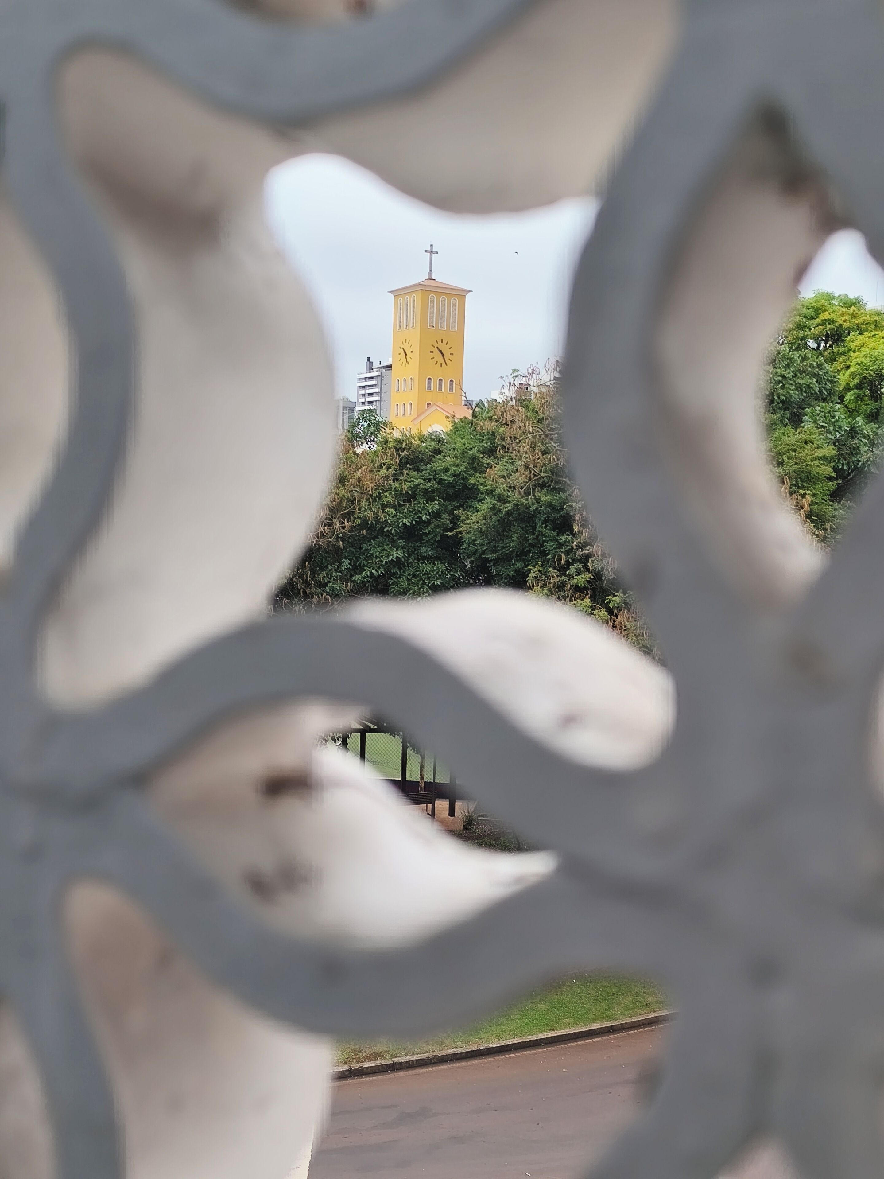 Un reloj de torre amarillo con una cruz en la parte superior, rodeado de vegetación.