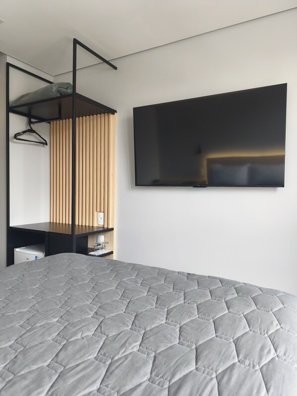 A modern hotel room with a large bed, a flat-screen TV mounted on the wall, a wooden slatted window, and a black hanging rack.