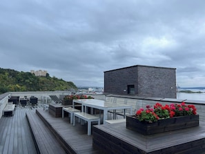 Una terraza en la azotea con cubierta de madera, mesas y bancos blancos, y macetas con flores.
