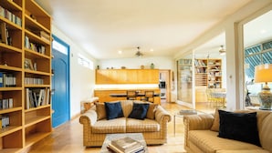 A spacious living area with a beige sofa, a coffee table with books, and a bookshelf filled with books. There is a kitchen area in the background with wooden cabinets and a dining table. - Family-Friendly Oasis with Pool (Cuernavaca)