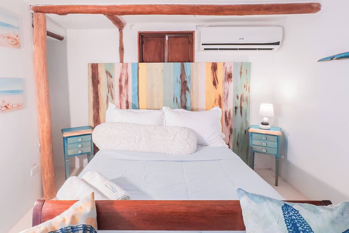 A bedroom with a wooden bed frame, white bedding, a colorful headboard, a small bedside table with a lamp, and a starfish pillow.