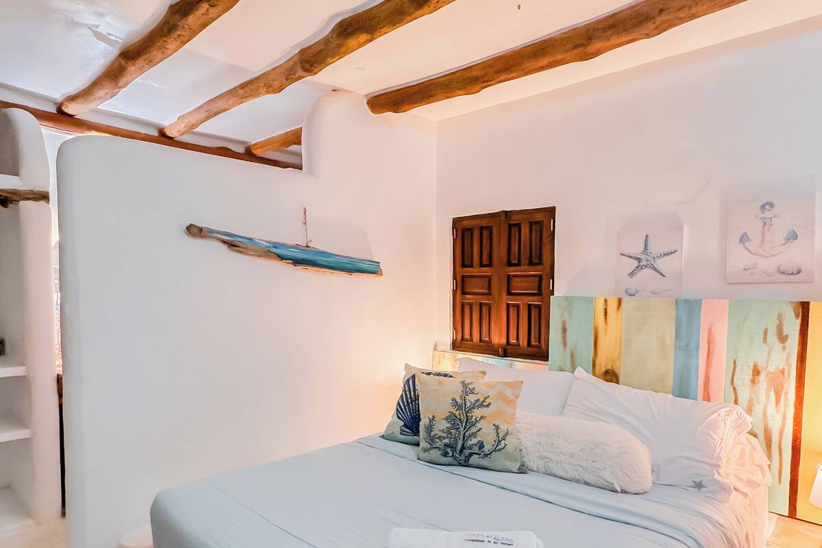 A bedroom with a bed, pillows, a wooden headboard, a small shelf, and a wall with nautical decorations.