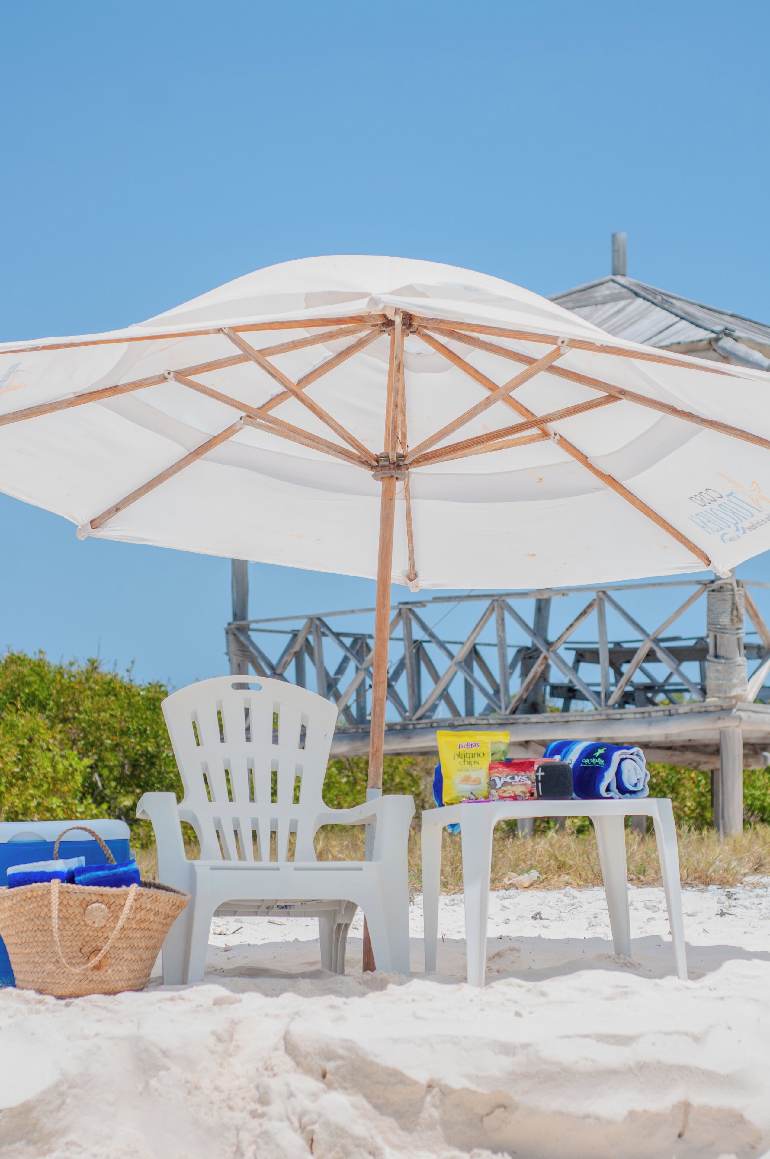 Een strandstoel onder een witte parasol, een koelbox en een stoel met lekkernijen op een tafel.