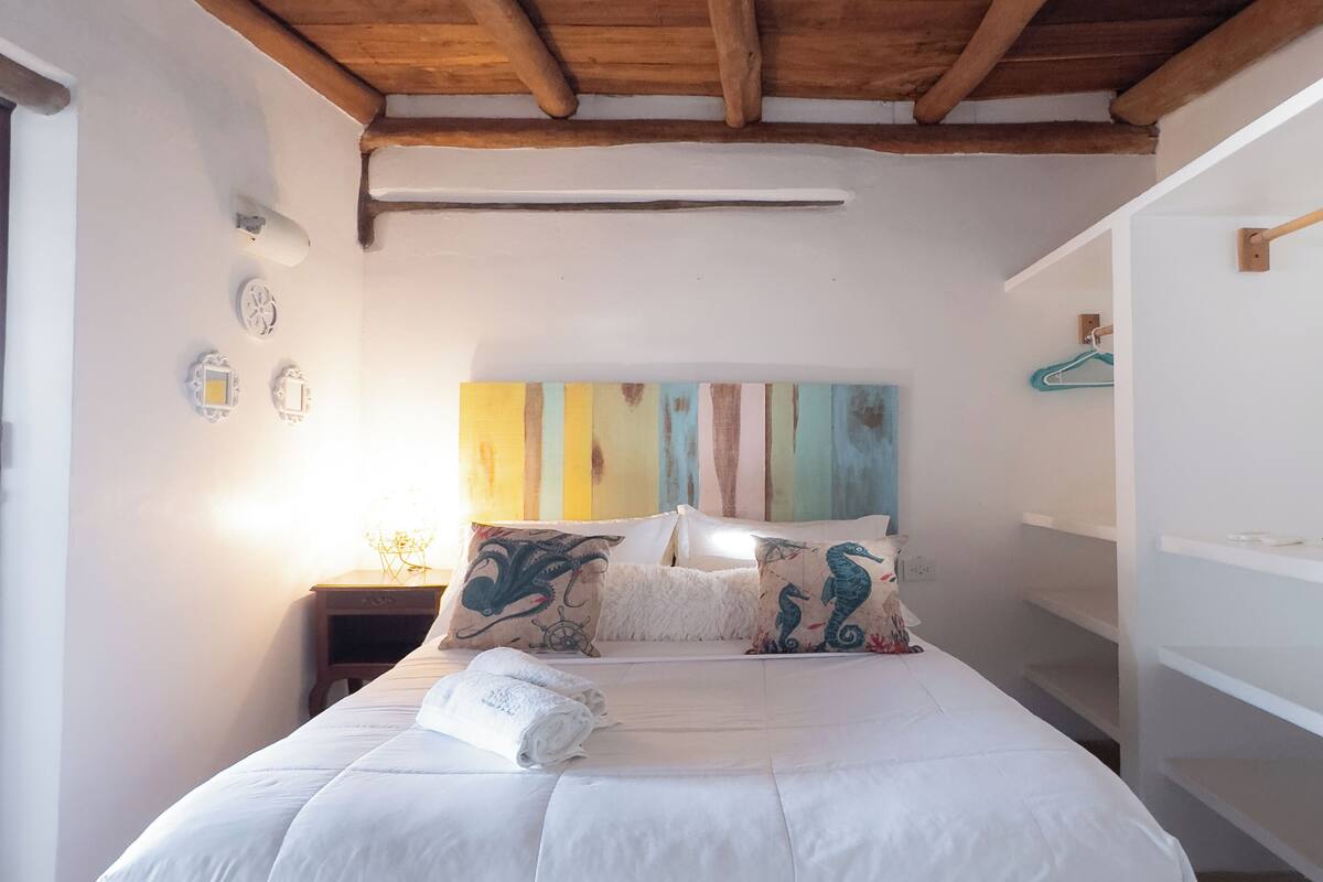 A bedroom with a wooden bed frame, white bedding, and a colorful wall art piece above the bed.