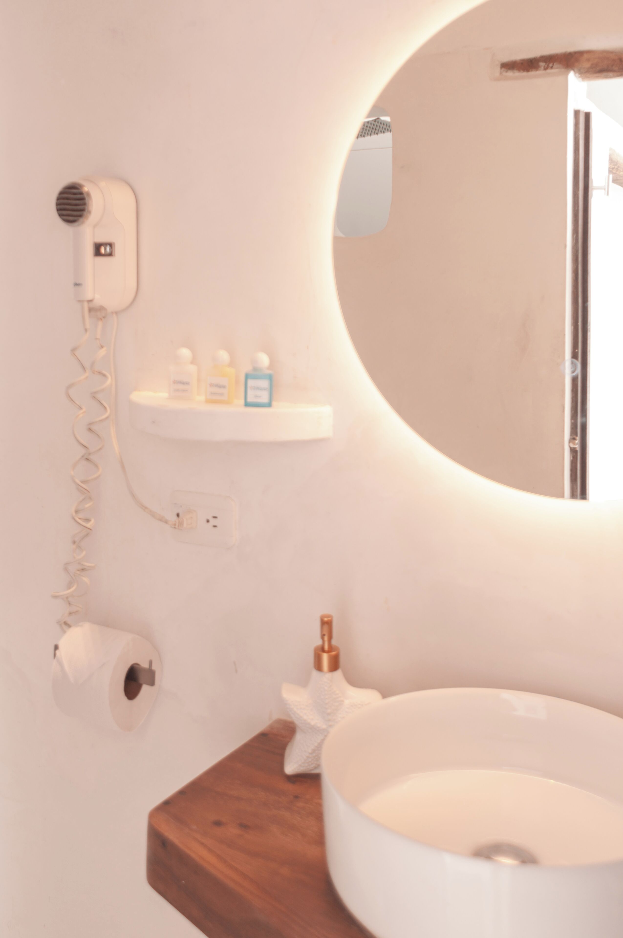 Un baño con un espejo redondo, un lavamanos blanco, un secador de pelo y un estante con artículos de aseo.