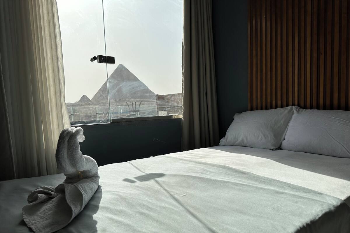 Quarto de hotel com cama, janela com cortinas e vista para uma pirâmide.