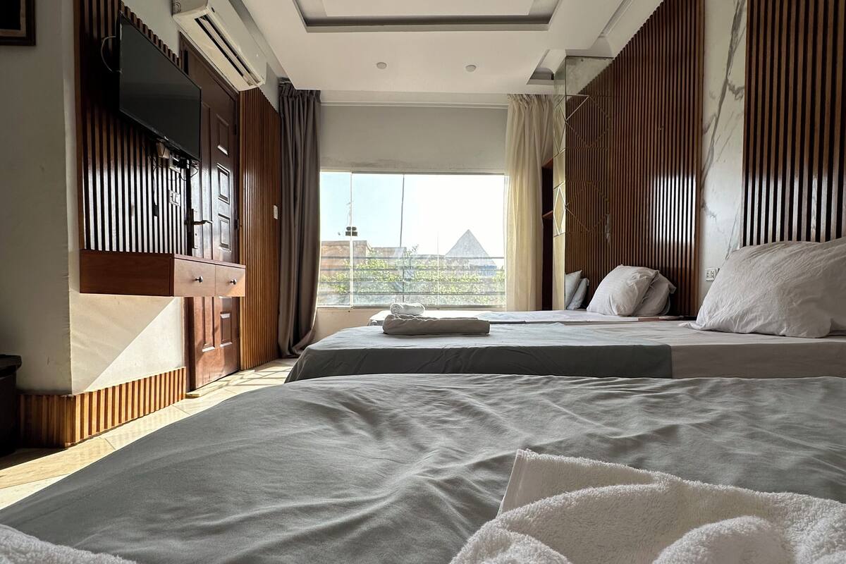 Um quarto moderno com uma cama grande, uma televisão fixada na parede e vista para a paisagem urbana pela janela.