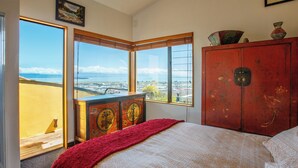 A bedroom with a large window offering a view of the city, a bed with a patterned bedspread, and a red cabinet with decorative items. - Sunset Heights - Nelson Holiday Home (Nelson)
