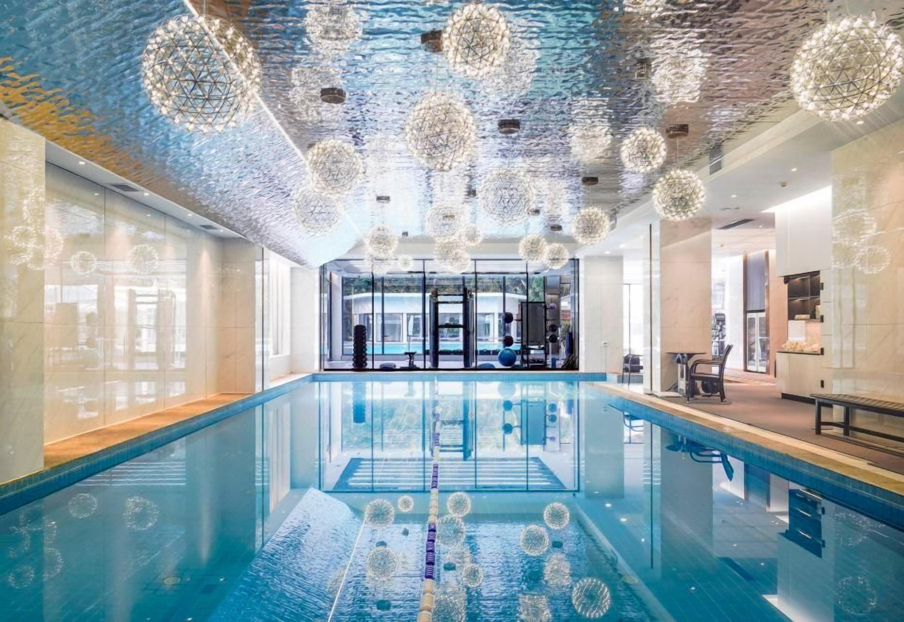 A modern indoor swimming pool with a glass barrier, surrounded by a spacious area with a high ceiling and decorative spherical light fixtures.
