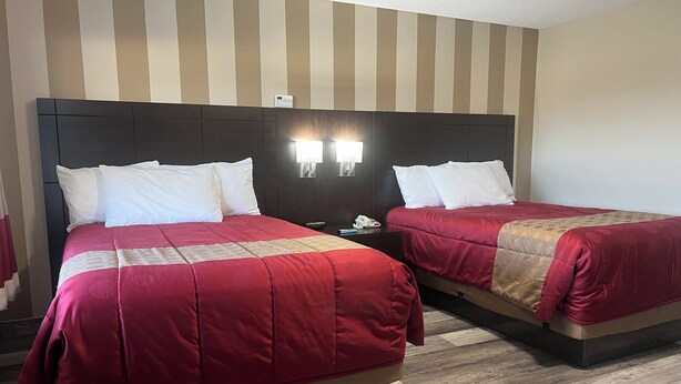 Ein Hotelzimmer mit zwei Betten, beide mit roter Bettdecke und weißen Kissen. Zwei Nachttische mit Lampen, einer davon mit einem Telefon. Die Wände sind mit Streifen versehen und der Boden ist mit Holz ausgelegt.