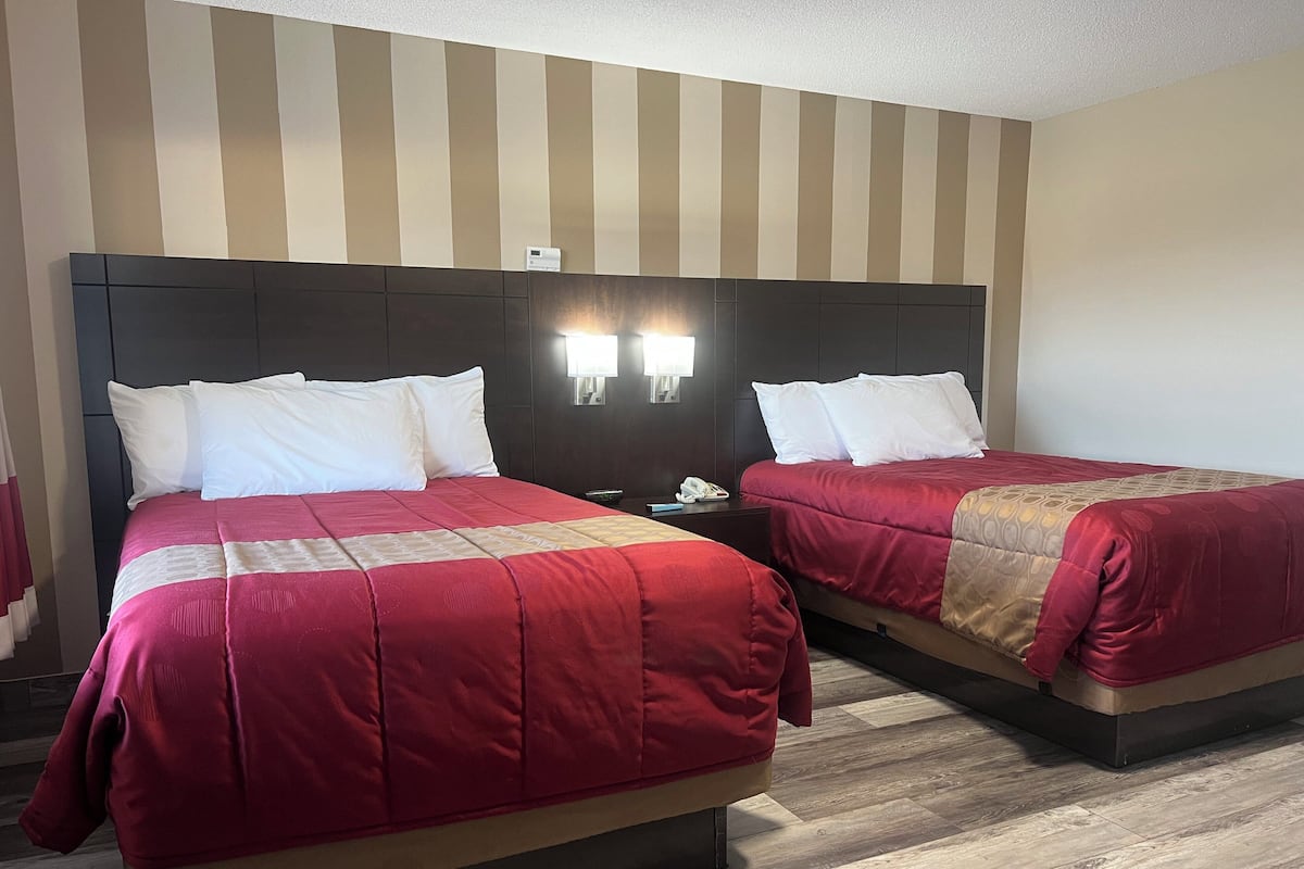 A hotel room with two beds, each with a red bedspread and white pillows. There are two bedside tables with lamps and a phone on one of them. The room has a striped wall design and wooden flooring.