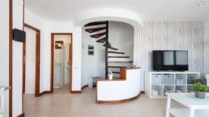 Un salon moderne avec un comptoir d’accueil blanc aux lignes courbes, un téléviseur noir posé sur un meuble blanc et un escalier en colimaçon menant à une autre pièce.