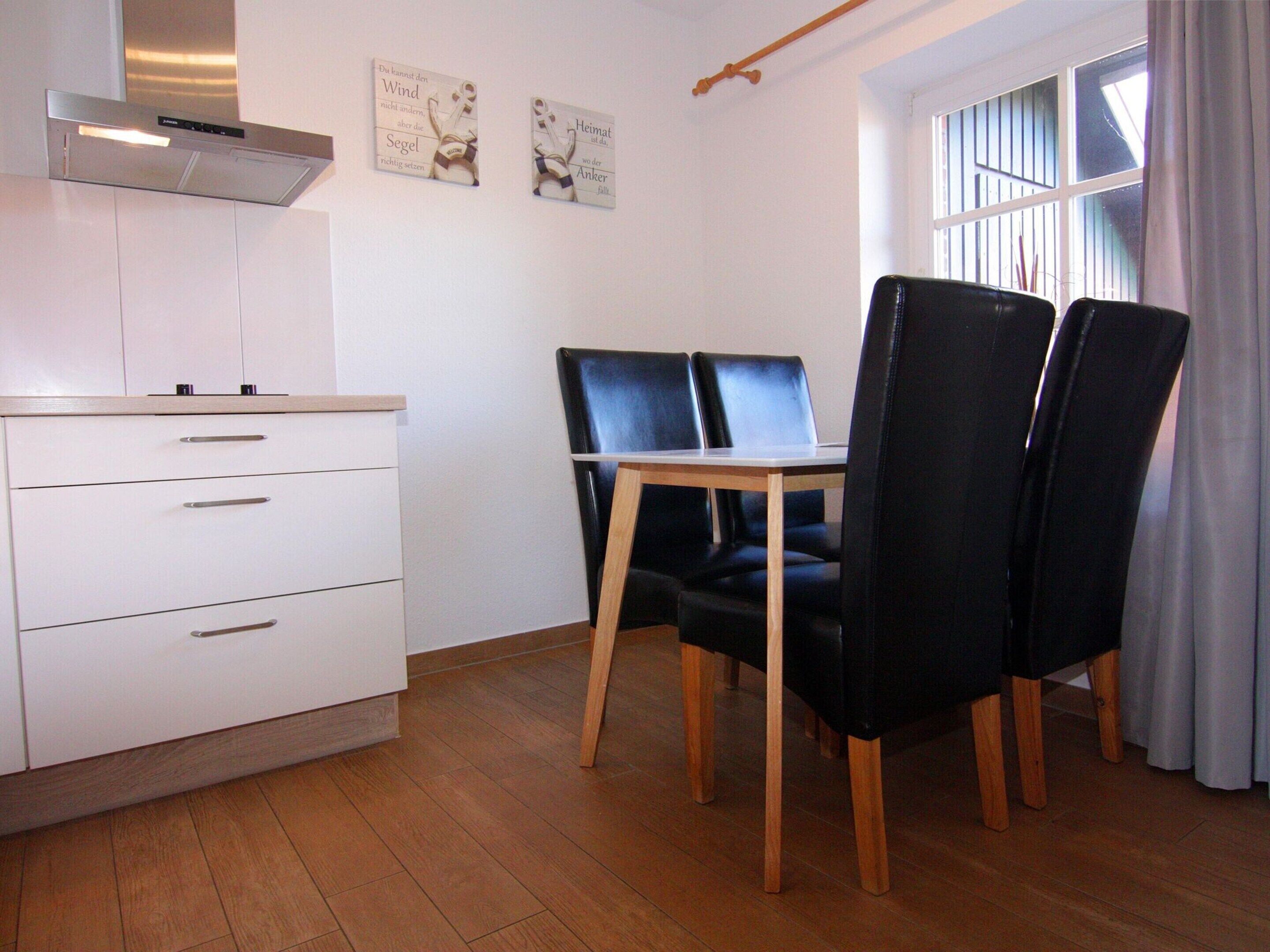 A modern kitchen with white cabinets and a stainless steel range hood. There is a dining table with black chairs and a window with blinds.