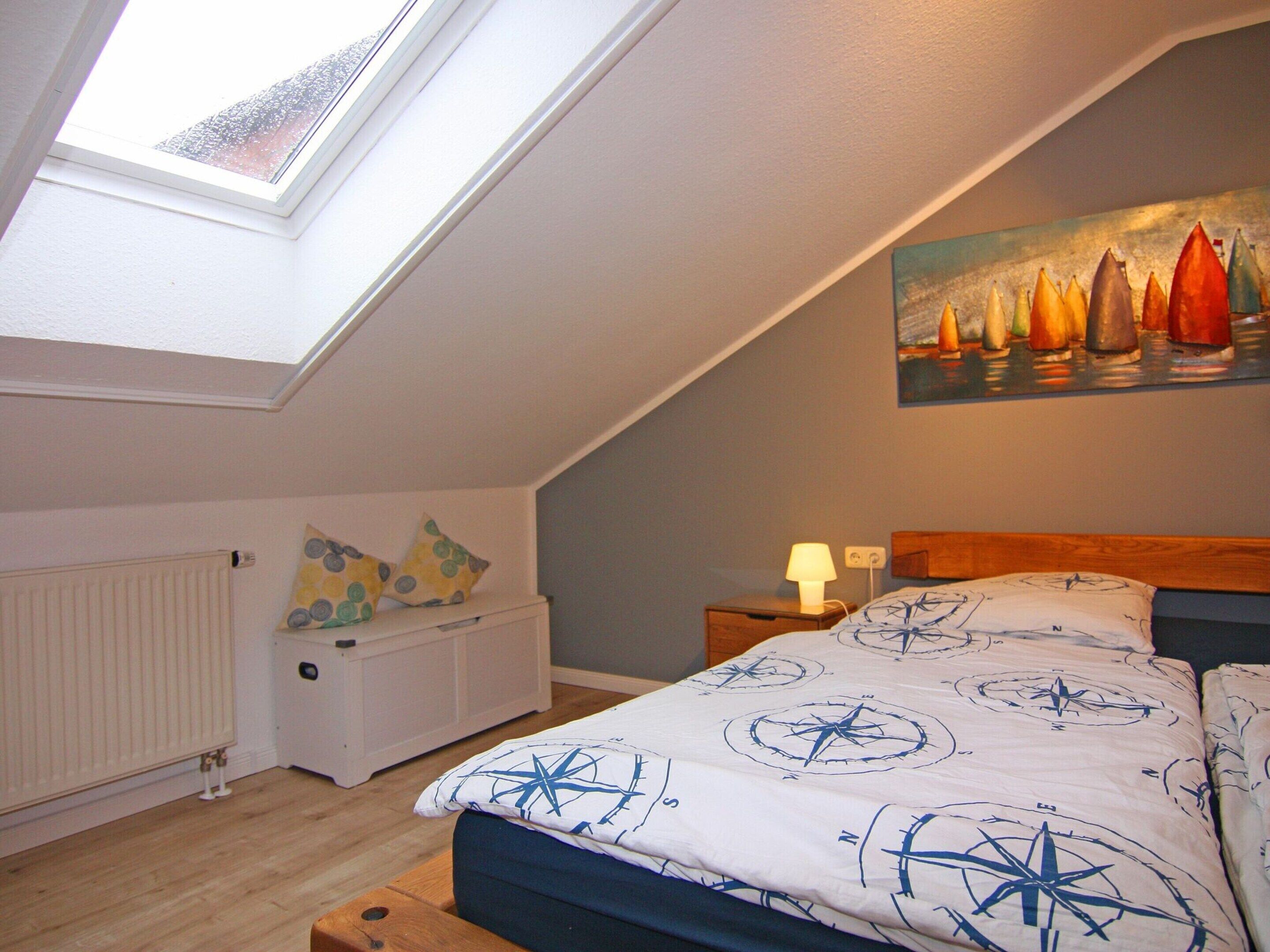 Ein Schlafzimmer mit Schrägwand, einem Dachfenster, einem Bett mit Kompassdecke, einem Nachttisch mit Lampe und einem Segelbootgemälde an der Wand.