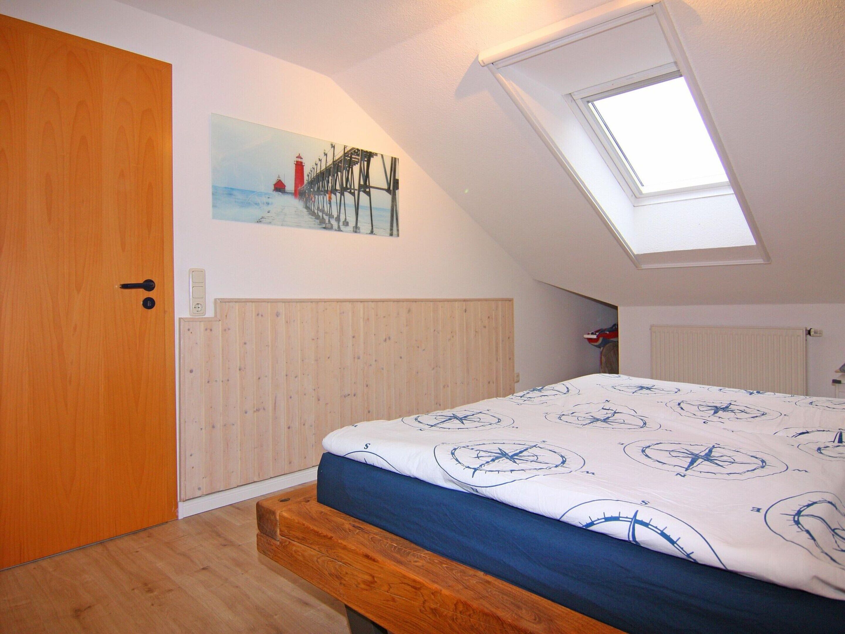 Une chambre avec un plafond en pente, un puits de lumière, un cadre de lit en bois, une tête de lit blanche et un tableau représentant un phare sur le mur.
