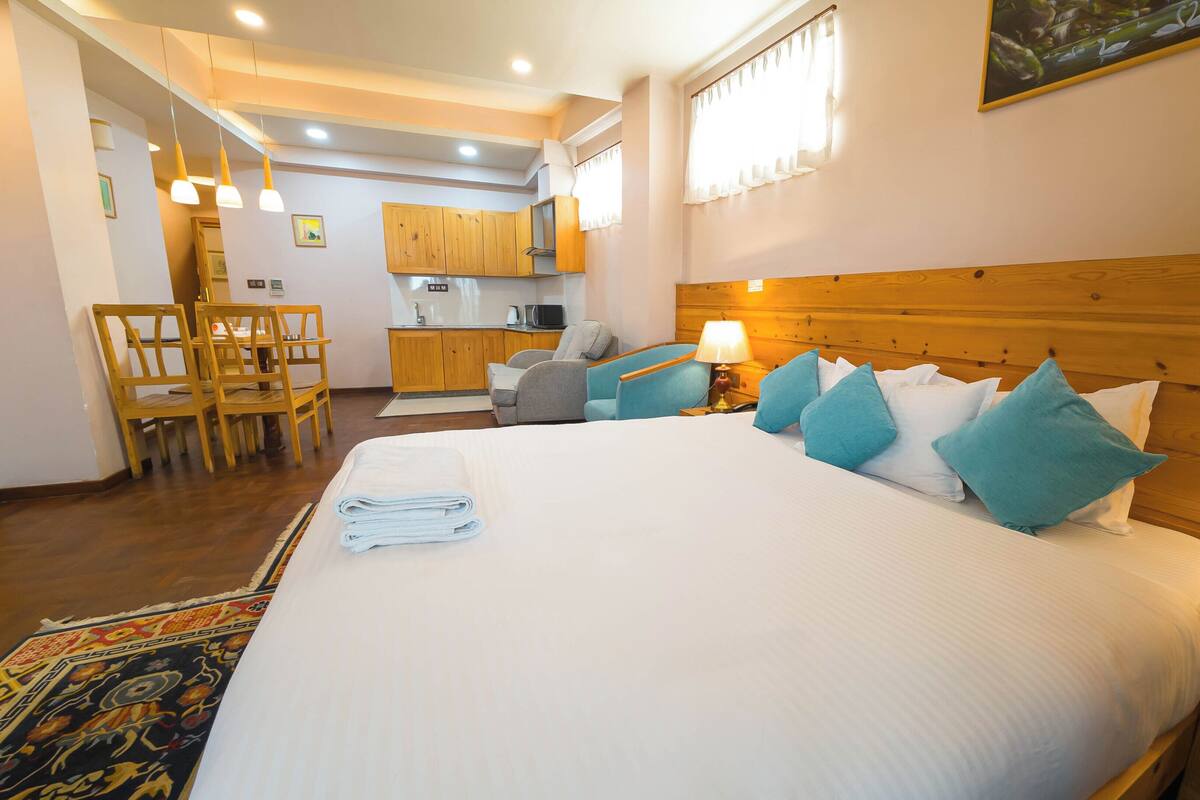 A hotel room with a large bed, a wooden headboard, a desk with a lamp, a chair, and a kitchenette in the background.