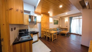 A modern kitchen with wooden cabinets, a microwave, and a sink. There is a dining area with a table and chairs, and a living room with a sofa and a lamp.