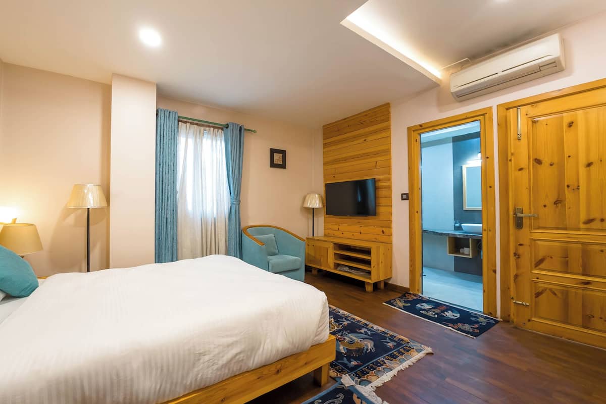 A bedroom with a bed, two bedside lamps, a TV, a wooden wardrobe, and a bathroom visible through an open door.