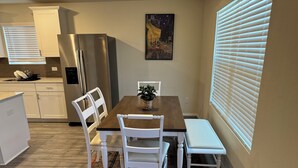 A dining area with a wooden table, white chairs, and a bench. A refrigerator is visible in the background.