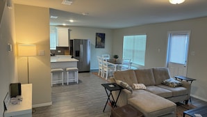 A modern living room with a sofa, ottoman, and a coffee table. There is a kitchen area with a refrigerator and a dining table with chairs.
