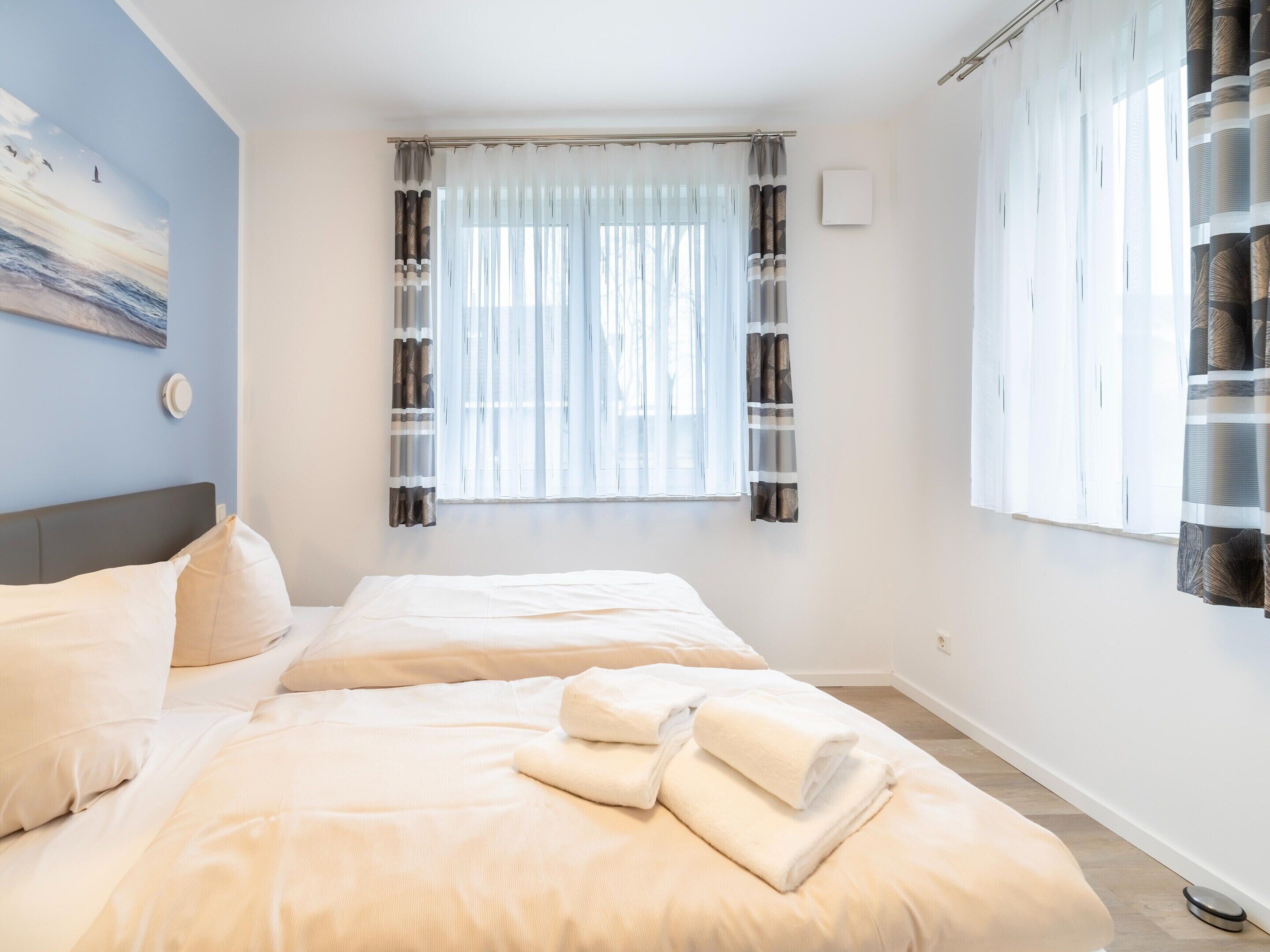 A bedroom with two beds, a window with sheer curtains, and a wall-mounted thermostat.