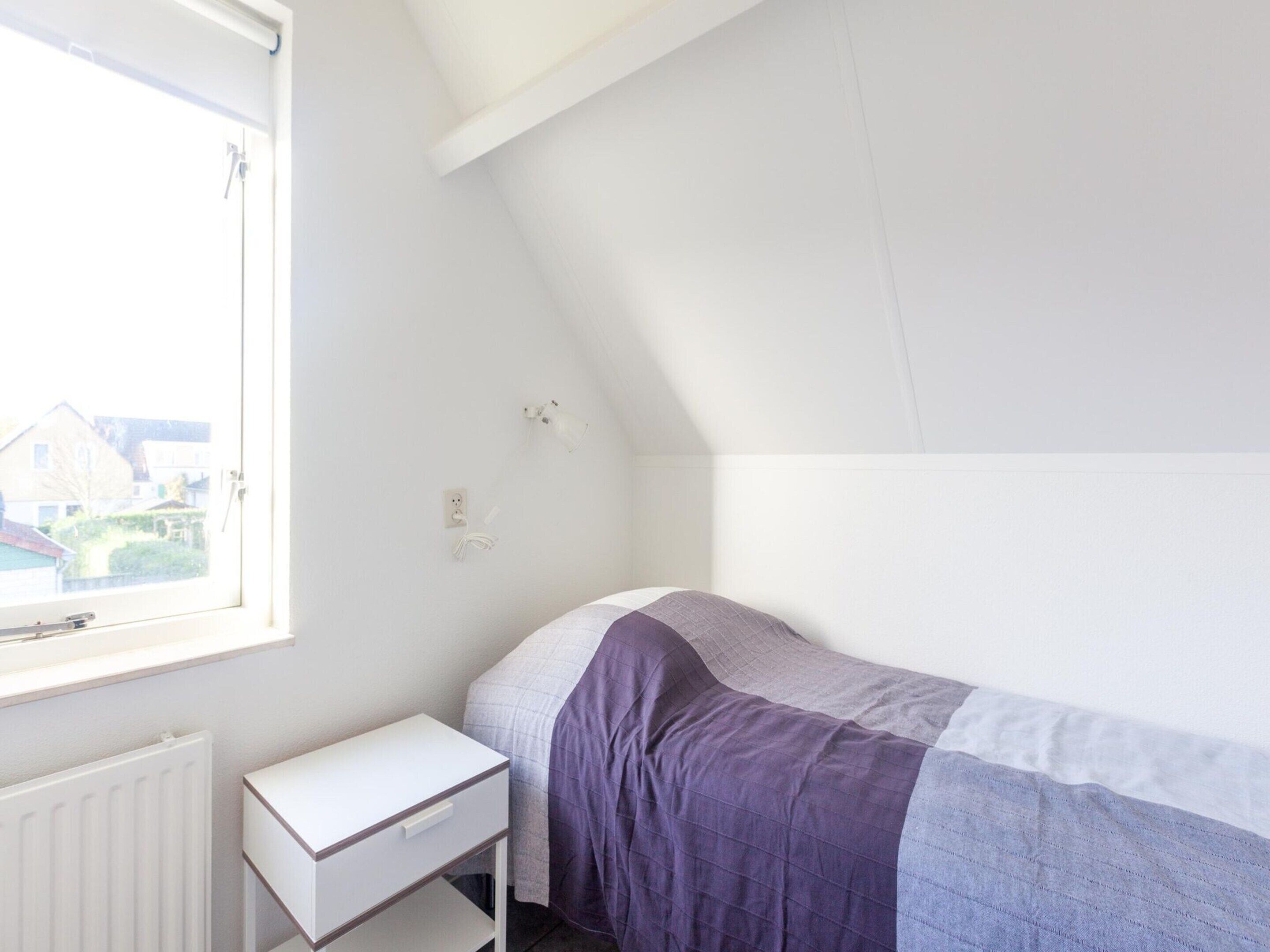 A small, simple room with a bed, a small white desk, and a window.
