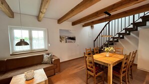 A dining area with a wooden table and chairs, a brown sofa, a window with a view, and a staircase. - Ferienwohnung auf Usedom im Lepels Hof bis zu 5 P (Luetow)