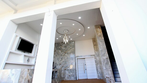 Un intérieur moderne avec un téléviseur à écran plat fixé à un mur blanc, un mur d’accent en marbre, un lustre et une porte blanche.