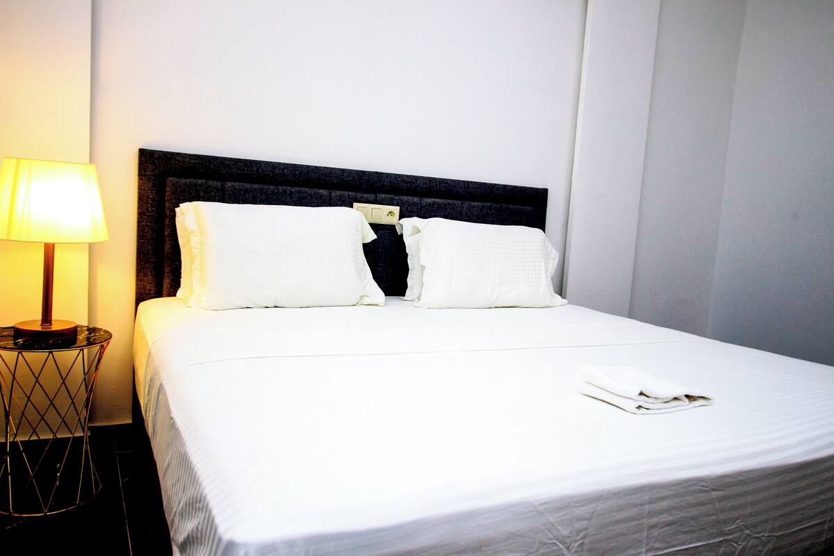 A neatly made bed with white linens and pillows, a bedside lamp, and a small round table with a geometric design.