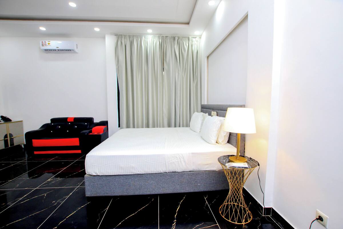 A hotel room with a bed, a desk, a chair, and an air conditioning unit.