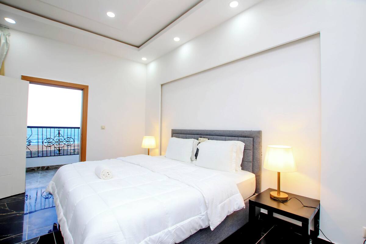 A hotel room with a bed, two bedside lamps, a view of the sea through a window, and a balcony with a railing.