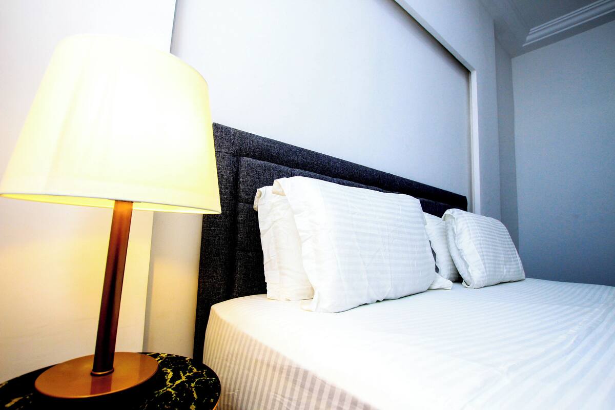 A neatly made bed with white linens and pillows, a bedside lamp, and a dark headboard.