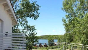 A wooden deck with a metal railing overlooking a lake, surrounded by trees and a few buildings in the distance. - Elegant Eingerichtete Ferienwohnung (Mirow)