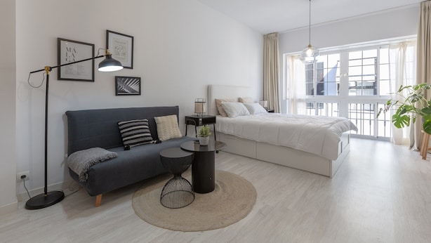 Un soggiorno moderno con un divano grigio scuro, un tavolino nero rotondo, un letto bianco e un'ampia finestra con tende.