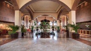 Un amplio vestíbulo con techos altos, columnas de arco y una lámpara central. Se observan varias plantas en macetas y un mostrador de recepción.