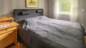 Ein Schlafzimmer mit einem Bett, einem Nachttisch, einer Lampe, einem gerahmten Bild an der Wand und einem Fenster mit Vorhängen.