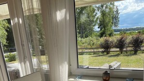 Ein Zimmer mit großem Fenster, das auf eine grüne Fläche mit Bäumen und einem Zaun blickt. Auf dem Fensterbrett steht eine Vase und zwei kleine Schalen.