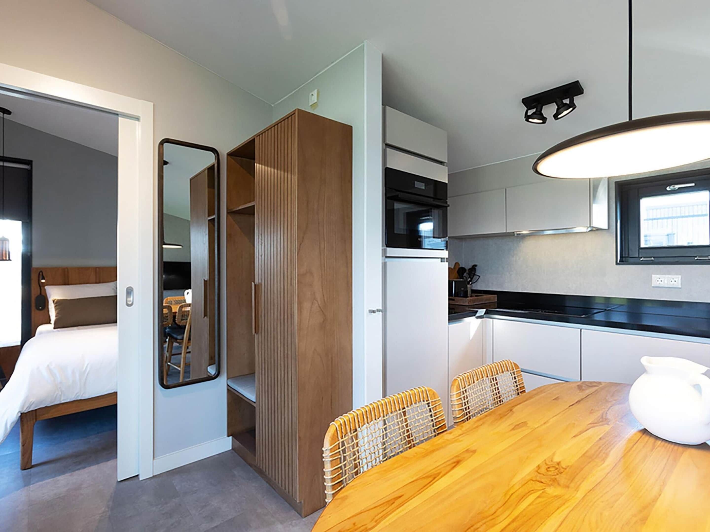 A compact hotel room with a wooden dining table, a kitchen area with white cabinets, and a large mirror.