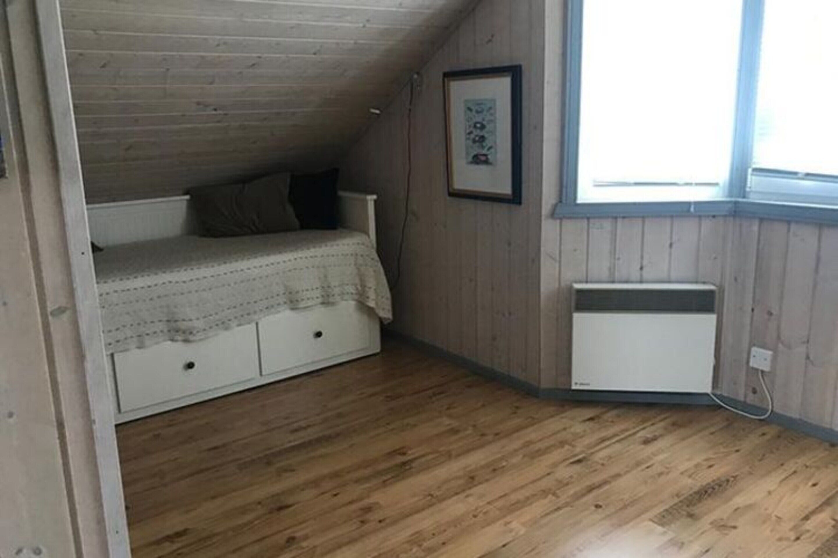 A small, single-room attic bedroom with a sloped ceiling, a bed with a patterned bedspread, a radiator, and a window providing natural light.
