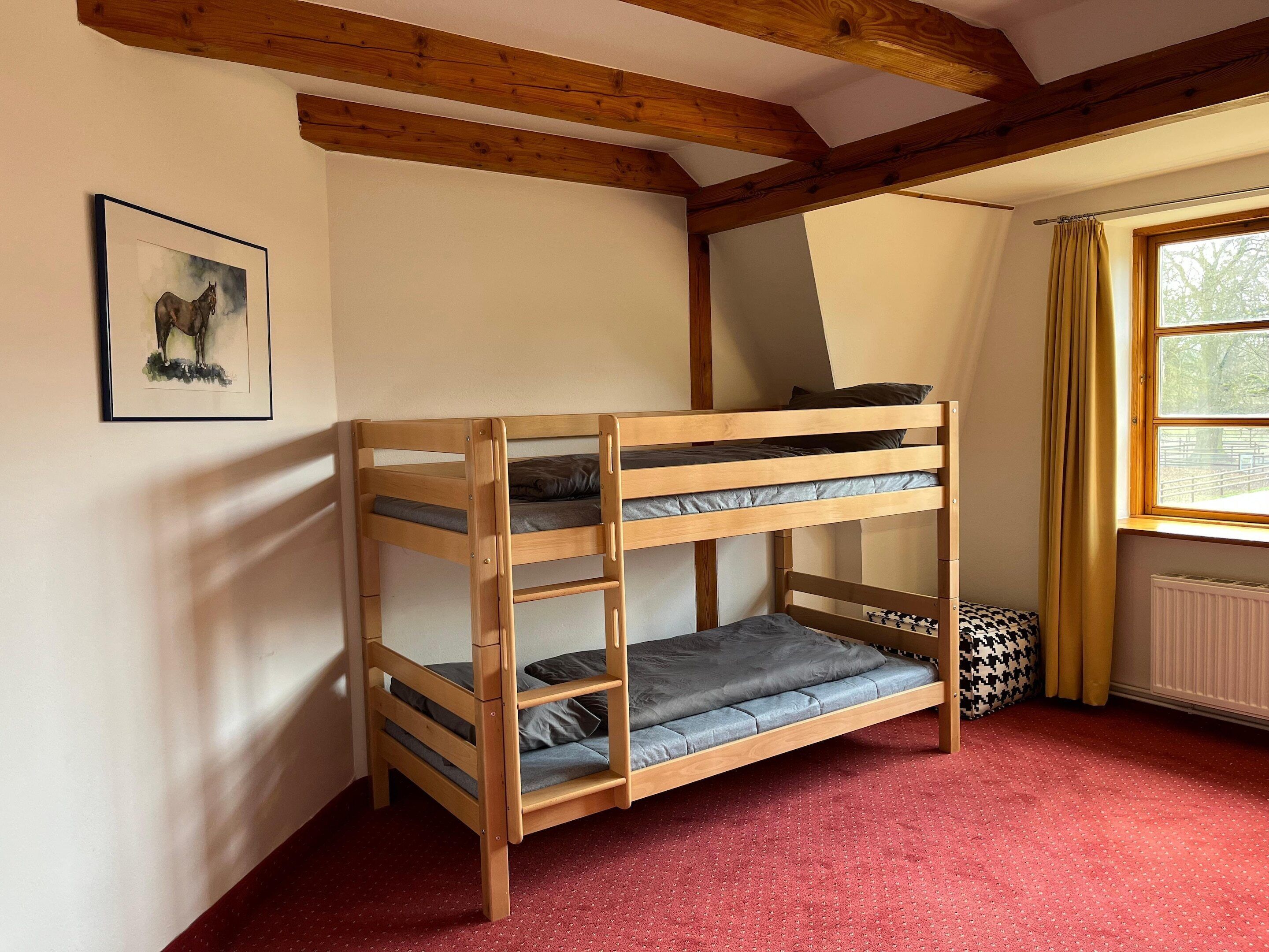 Una cama individual con estructura de madera y ropa de cama gris, un cuadro en la pared y una ventana con cortinas.