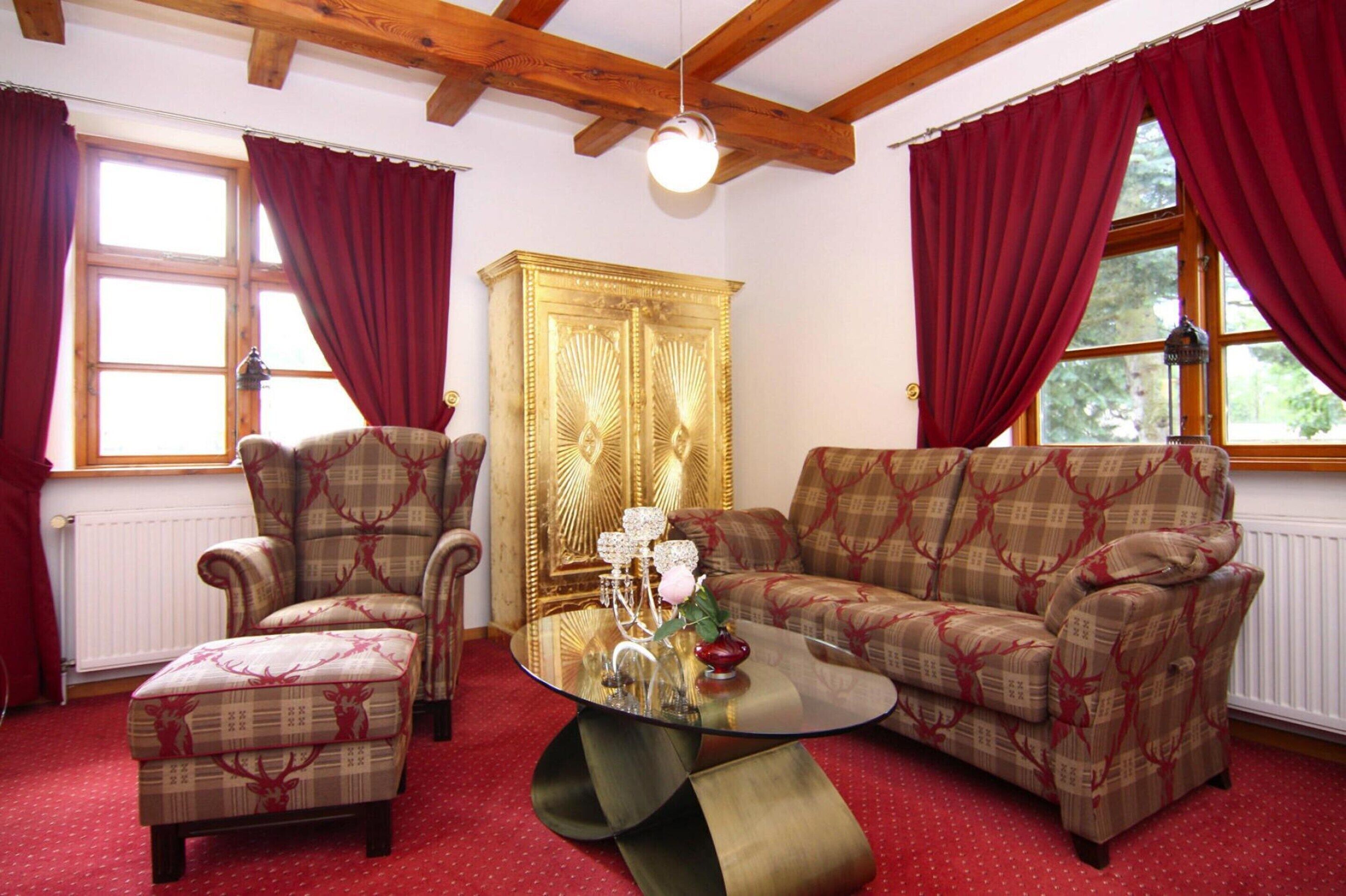 A room with a wooden ceiling, red curtains, a patterned sofa, a matching armchair, and a glass coffee table with a vase and candle.