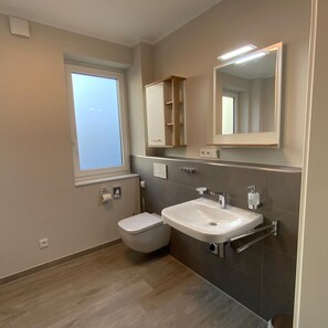 Une salle de bain moderne équipée de toilettes, d’un lavabo et d’un miroir. Une fenêtre laisse entrer la lumière naturelle et une étagère intégrée se trouve au-dessus du lavabo.