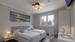 A modern bedroom with two beds, a desk, a chair, and a large window with a view of a building. - Exclusives Appartement auf Fehmarn (Fehmarn)